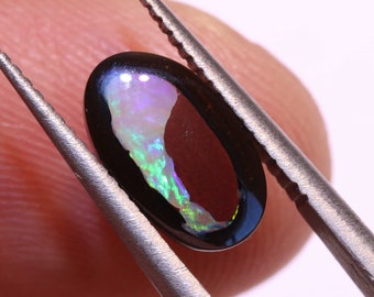 Pode incluir: Uma pedra preciosa de opala negra em forma oval, segurada por uma pinça. A opala exibe um vibrante jogo de cores, com flashes de verde, azul e roxo em seu interior. A pedra preciosa é polida e tem uma superfície lisa.