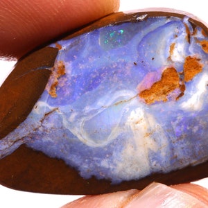 May include: A polished opal gemstone with a dark brown outer layer and a vibrant interior. The interior displays swirling patterns of blue, white, and hints of pink and green. The stone is held by a finger.