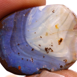 May include: A polished, irregular-shaped boulder opal gemstone featuring a vibrant blue section transitioning into a translucent, milky white area. The surface displays natural brown matrix inclusions and subtle, swirling color patterns throughout.