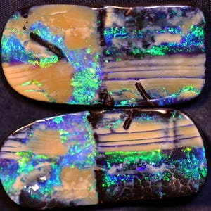 Könnte beinhalten: Zwei Stücke Opal mit schwarzer Matrix und schillernden grünen, blauen und violetten Farben. Der Opal hat ein gestreiftes Muster.