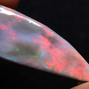 May include: A polished opal gemstone with a pearlescent white base, displaying vibrant internal colors of red, green, and blue. The stone has a smooth, reflective surface and a unique, elongated shape, perfect for jewelry.