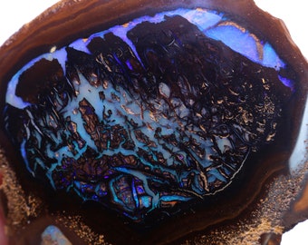 Pode incluir: Uma pedra de opala polida com uma camada externa marrom escura e um interior vibrante. O interior apresenta uma mistura de tons azuis, verde-azulados e roxos com padrões intrincados. A pedra tem uma forma orgânica única.