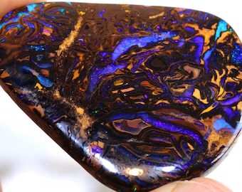 Pode incluir: Um opala marrom escuro, azul e roxo com um padrão de redemoinho. O opala é um cabochão, uma gema lisa e arredondada.
