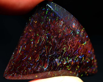 May include: A triangular-shaped gemstone with a dark red base color and intricate, iridescent patterns. The patterns display a range of colors, including green, blue, and yellow, creating a shimmering effect. The stone is held by a person's fingers.