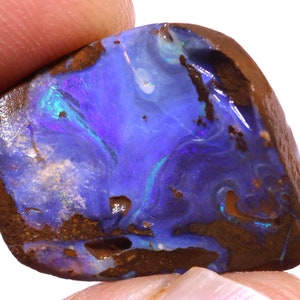 Könnte beinhalten: Ein polierter Opal-Edelstein mit einer lebendigen Darstellung von blauen, violetten und grünen Farben. Der Opal hat eine dunkelbraune Matrix und eine glatte, glänzende Oberfläche. Der Edelstein wird zwischen zwei Fingern gehalten.