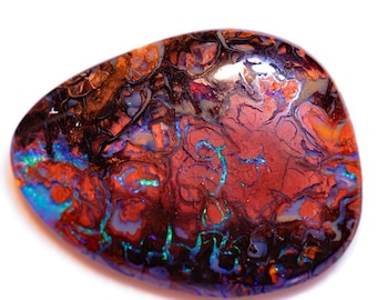Puede incluir: Una gema pulida de forma ovalada con una vibrante exhibición de colores. La piedra presenta una mezcla de tonos rojos, naranjas, azules y verdes, con patrones oscuros e intrincados. La gema se encuentra sobre un fondo blanco.