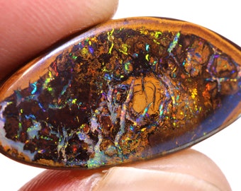 Pode incluir: Um close-up de um opala marrom e iridescente com um padrão único de veios e redemoinhos. O opala está configurado em uma forma de cabochão e tem uma superfície lisa e polida.
