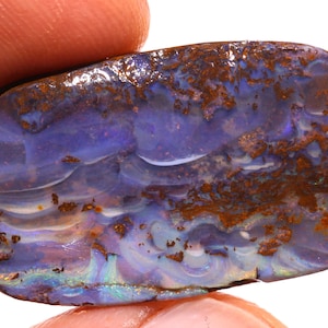 May include: A polished, rectangular opal gemstone with a swirling pattern of blues, purples, and whites. The surface has areas of iridescent color and is framed by a band of brown matrix.