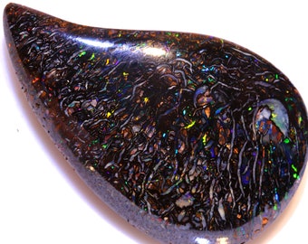 Pode incluir: Um opala preto em forma de lágrima com uma superfície escura e iridescente. O opala tem um padrão complexo de veios e redemoinhos em tons de azul, verde e dourado.