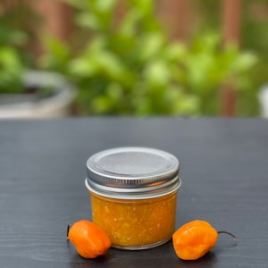 May include: A small glass jar filled with a bright orange sauce, with a silver lid. Two small orange habanero peppers are on either side of the jar.