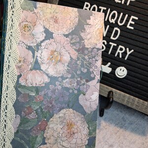 May include: A floral journal with a cream lace spine. The cover features a watercolor design of pink and peach flowers with green leaves on a blue background. The words "TULIP BOTIQUE AND POETRY" are on a letter board.