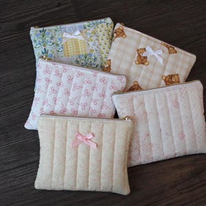 May include: Five quilted fabric pouches in various patterns and colors. One pouch has a floral pattern, another has teddy bears, and a third has pink bows. Each pouch has a zipper closure and a bow accent.