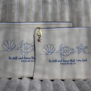 May include: Two off-white canvas zipper pouches featuring blue embroidered line art of a sea turtle, a seashell, and a starfish. The text "Be still and know that I am God PSALM 46:10" is stitched below the designs. Each zipper has a gold-toned charm.