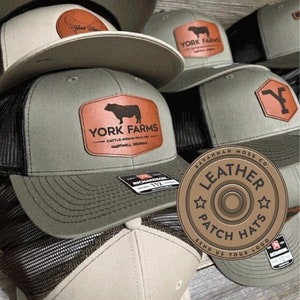 May include: A stack of green and beige baseball caps with leather patches. The patches feature a bull, a Y, and the text "YORK FARMS" and "SAVANNAH MOSS CO. LEATHER PATCH HATS SEND US YOUR LOGO".