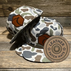 May include: Two camouflage patterned baseball caps with a brown leather patch on each. The patch on the front cap has a stylized letter 'A' and the patch on the back cap has a stylized peach. The back cap also has a brown leather circle with the text "Savannah Moss Co. Leather Patch Hats Send Us Your Logo" in white.