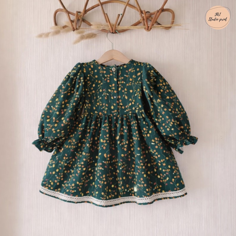 May include: A dark green dress with a floral pattern of yellow and orange dots. The dress has long sleeves with ruffled cuffs, a round neckline, and a white lace trim at the hem. The dress is hanging on a wooden hanger.