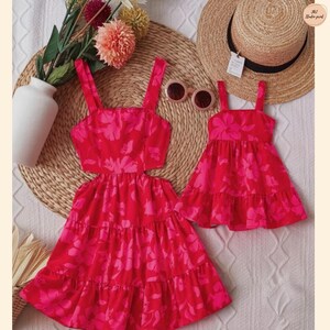 May include: Two matching red dresses with a floral pattern. One dress has a cutout design, while the other is a smaller version. The dresses are placed on a woven mat with a straw hat and sunglasses.