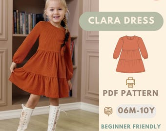 May include: A young girl wearing an orange Clara dress with long sleeves and a tiered skirt. The image includes a PDF pattern graphic, text that reads "CLARA DRESS", and "BONUS: APRON PATTERN". The dress is available in sizes 06M-10Y.