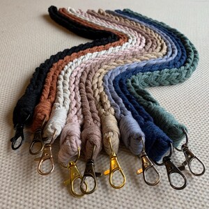 May include: A collection of braided macrame lanyards arranged in a wavy pattern. The lanyards feature various colours including black, rust, white, pink, beige, tan, light blue, navy, and sage green, each finished with a metal lobster clasp.