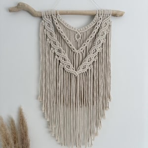 May include: A beige macrame wall hanging suspended from a natural light-coloured wooden branch. The piece features intricate knotted patterns arranged in two V-shaped layers, with long, loose fringe hanging down to form a pointed bottom edge.
