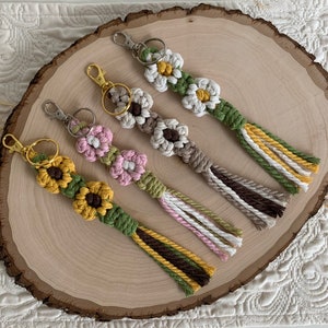 May include: Four handmade macrame keychains with flower designs. The keychains feature a variety of colours, including green, yellow, brown, white, pink, and beige. Each keychain has a tassel at the end.