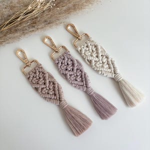 May include: Three macrame keychains in beige, lavender, and cream. Each has a woven design, a tassel, and a gold-coloured clasp. The keychains are arranged diagonally against a white background, with dried ornamental grass in the background.