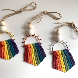 May include: Three rainbow-coloured macrame wall hangings with wooden beads and metal hoops. Each hanging has a different coloured hoop: gold, copper, and silver.