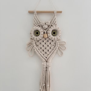 May include: A beige macrame owl wall hanging. The owl has large, circular eyes with green centres and a wooden bead for a beak. The owl is suspended from a wooden dowel and has a tassel at the bottom. The owl is made of knotted cotton rope.