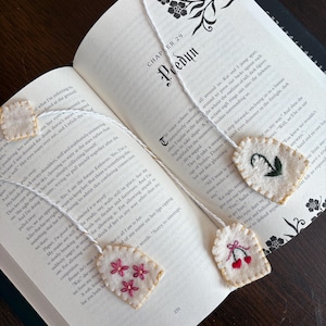 May include: Open book with several handmade felt bookmarks. The bookmarks are white with embroidered floral and cherry designs. The book is open to chapter 29, with text visible on the pages. The bookmarks are attached to the pages with white string.