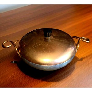 May include: A vintage copper-colored cooking pot with a lid and two loop handles. The pot has a silver-colored base and a copper-colored lid with a black knob. The handles are gold-colored. The pot is sitting on a wooden surface.