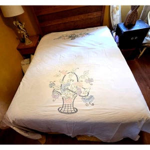 May include: White bedspread with an embroidered floral basket design. The basket is filled with pink, blue, and purple flowers and green leaves. The bedspread is on a bed with a wooden headboard.
