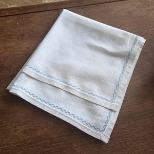 May include: Folded white linen napkin with a light blue embroidered border. The napkin is neatly folded and appears to be made of a soft, natural fabric. The blue embroidery adds a delicate touch to the classic design.