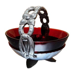 May include: A decorative bowl with a ruby red interior and a silver-colored exterior. The bowl has a handle with a floral design and rests on three feet. The handle is adorned with flower-shaped details.