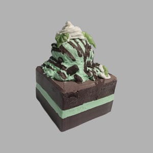 May include: A square-shaped dessert featuring layers of chocolate and mint, topped with mint ice cream, chocolate drizzle, whipped cream, and green leaf accents. The dessert is a miniature replica of a mint chocolate dessert.