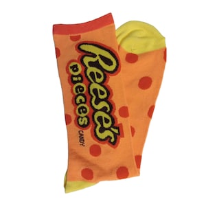 May include: A pair of orange socks with red polka dots and the "Reese's Pieces Candy" logo in black and yellow. The toe and cuff are yellow and orange, respectively. These socks are a fun and colorful accessory.