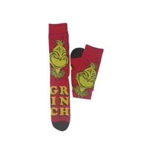 May include: Red socks featuring a yellow Grinch face and the word "GRINCH" in yellow block letters. The socks have black heels and toes. The Grinch's face is on the front of the socks.