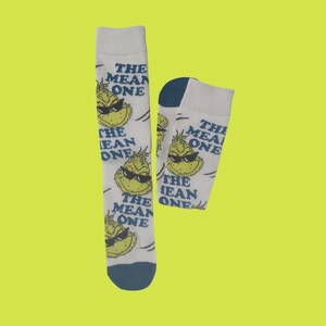 May include: A pair of white socks with a repeating pattern of a green cartoon character wearing sunglasses and the text "THE MEAN ONE." The socks have blue accents on the toe and heel.