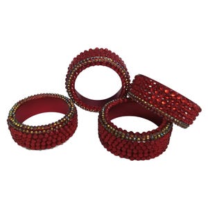 May include: A set of four circular napkin rings in a deep red hue. Each ring is densely covered in small, faceted red beads and features a thin, shimmering border of iridescent, multi-colored crystals along the top and bottom edges.