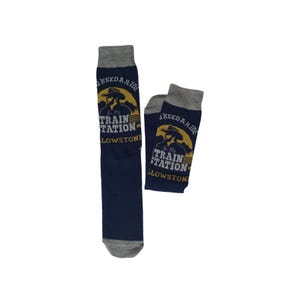 May include: A pair of navy blue socks with grey cuffs and toe sections. The socks feature a yellow graphic of a person in a cowboy hat with the text "NO NEED A RIDE TO THE TRAIN STATION" and "YELLOWSTONE" printed in white and yellow lettering.