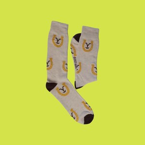 May include: A pair of light gray crew socks featuring a repeating pattern of yellow horseshoe icons. The socks have dark brown heels and toes. The text 2025 spike is printed in small black letters along the side of one of the socks.