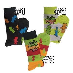 May include: Three pairs of socks with colorful designs. One pair is black with Sour Patch Kids characters in various colors. Another pair is white with green stripes and Sour Patch Kids graphics. The third pair has a striped pattern with the Sour Patch Kids logo.