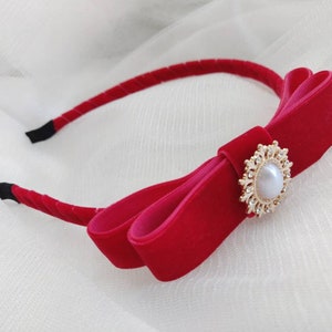 Puede incluir: Una diadema de terciopelo rojo con un gran lazo dorado y un botón nacarado en el centro. La diadema está sobre un fondo blanco.