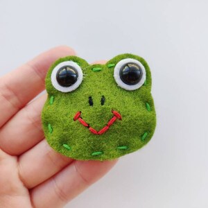 Op de afbeelding: Een groene vilt kikker met witte ogen en een rode gestikte glimlach.
