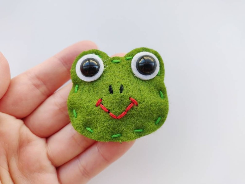 Op de afbeelding: Een groene vilt kikker met witte ogen en een rode gestikte glimlach.