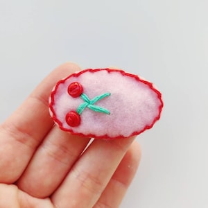 Op de afbeelding: Een roze ovaal van vilt met een rode gestikte rand en twee rode geborduurde kersen met groene stelen.