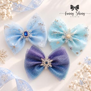 Puede incluir: Tres lazos para el pelo hechos de tul transparente azul y morado con patrones de copos de nieve plateados. Cada lazo presenta un adorno central de copo de nieve con una piedra azul o blanca. El texto "funny Shiny STUDIO" aparece arriba.