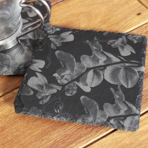 May include: Black slate coasters with a gray orchid design. The coasters are square and have a rough, natural edge.