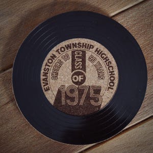 May include: A black vinyl record coaster with a cork center. The cork center has a brown inscription that reads "Evanston Township High School Cheers to Class of 1975 50 Years".