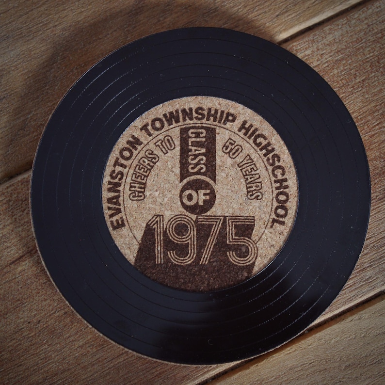 May include: A black vinyl record coaster with a cork center. The cork center has a brown inscription that reads "Evanston Township High School Cheers to Class of 1975 50 Years".