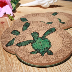 May include: Four round cork coasters featuring a green sea turtle design. The turtles are surrounded by a swirling pattern.